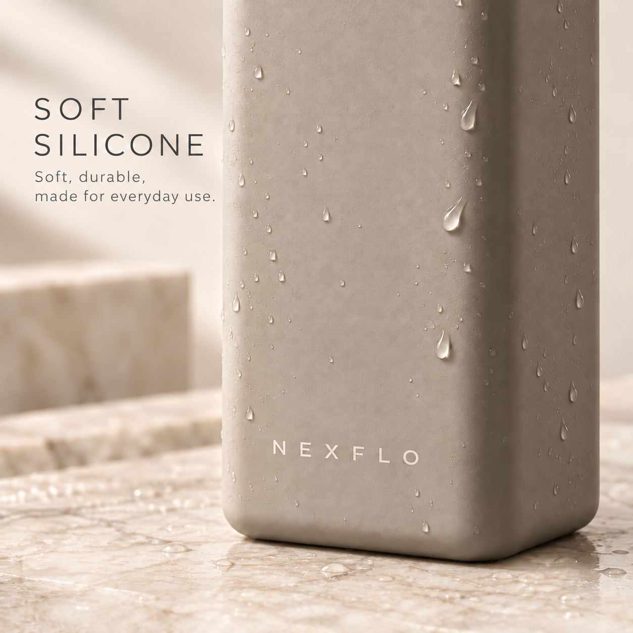 Soft silicone. Soft, durable, made for everyday use. Stone Grey NEXFLO square bottle close-up with water droplets on a warm marble pedestal.